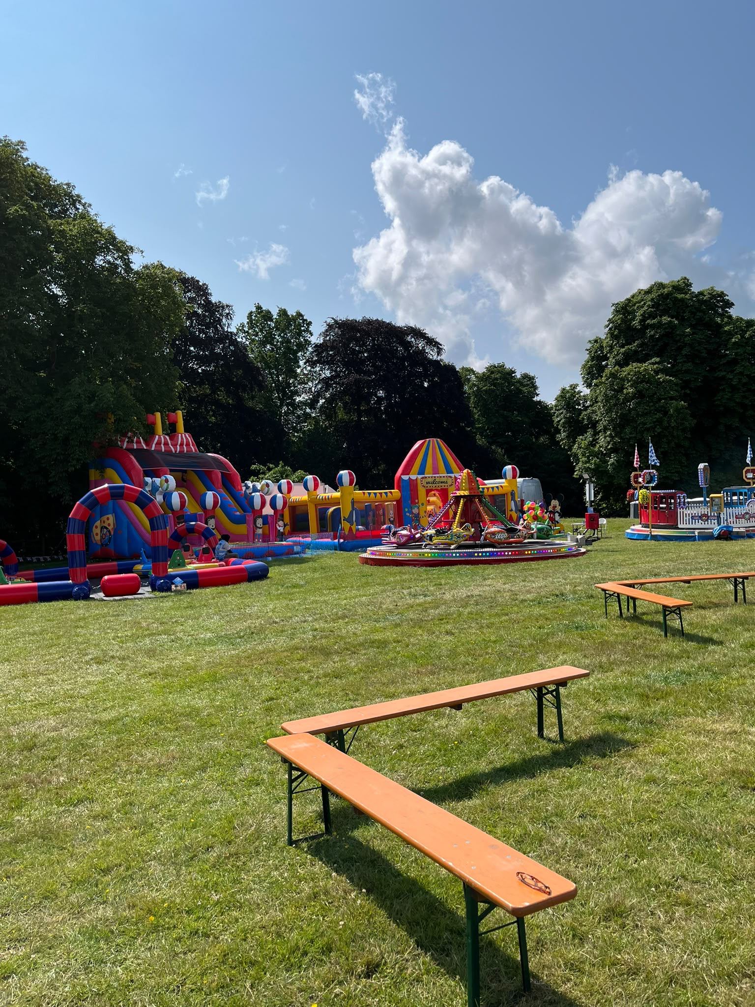 A wide panorama of a community fun day with a slide, welcome arch inflatable, chairoplane ride and a Toy Town train ride